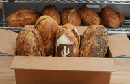Four large loaves of bread in a box. One has seeds encrusted in the crust, one has raisins visible, one looks plain, and one has an image of a saguaro made from a dusting of  flour.